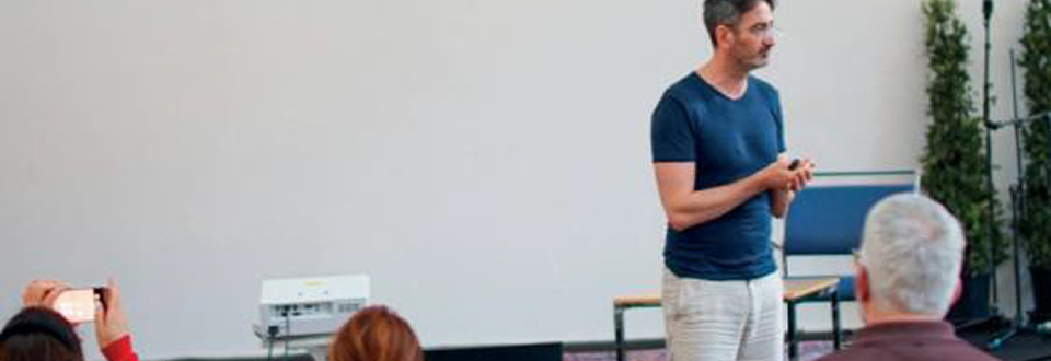 Personne debout devant un public, donnant une présentation ou un exposé dans une salle, avec un mur blanc derrière lui et quelques chaises visibles, tandis que des spectateurs sont assis et écoutent.