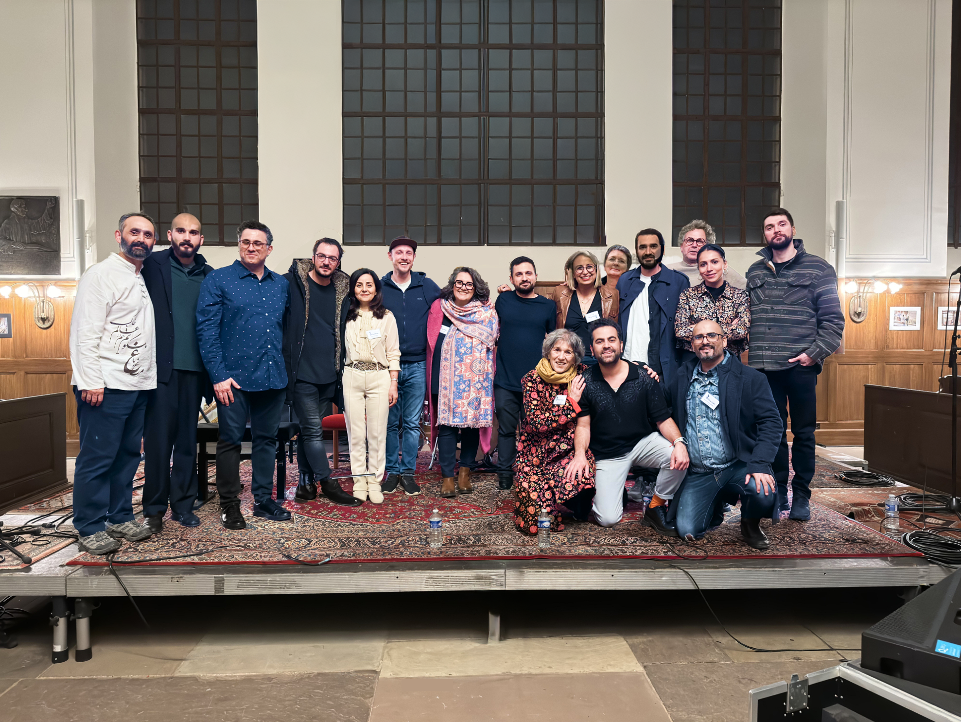 Groupe de personnes posant ensemble sur une scène ou une estrade devant une grande fenêtre intérieure, debout ou accroupies, souriantes, formant une photo de groupe prise à la fin d’un événement culturel